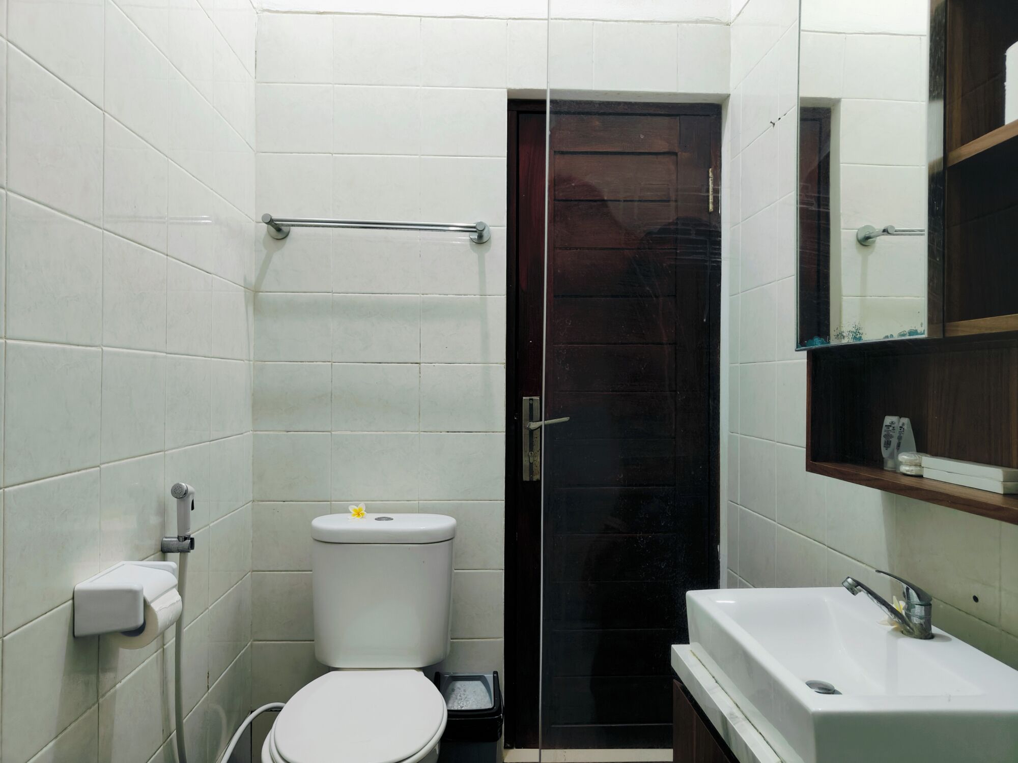 A small bathroom with white tiled walls, a white toilet, a black door, a glass shower enclosure, and a white sink with a mirror above it.