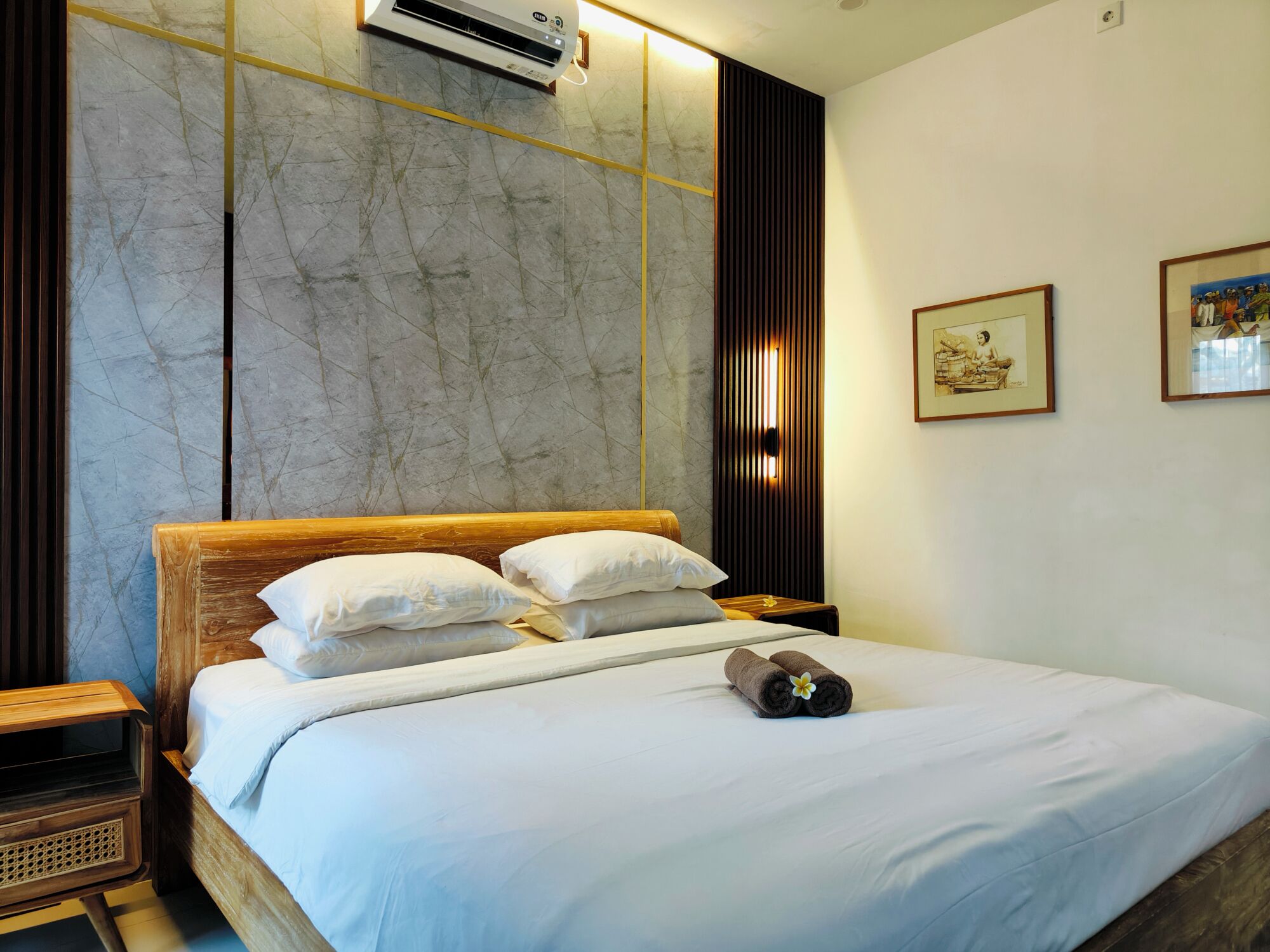 A hotel room with a large bed, a wooden headboard, a wall-mounted air conditioner, and two framed artworks on the wall.