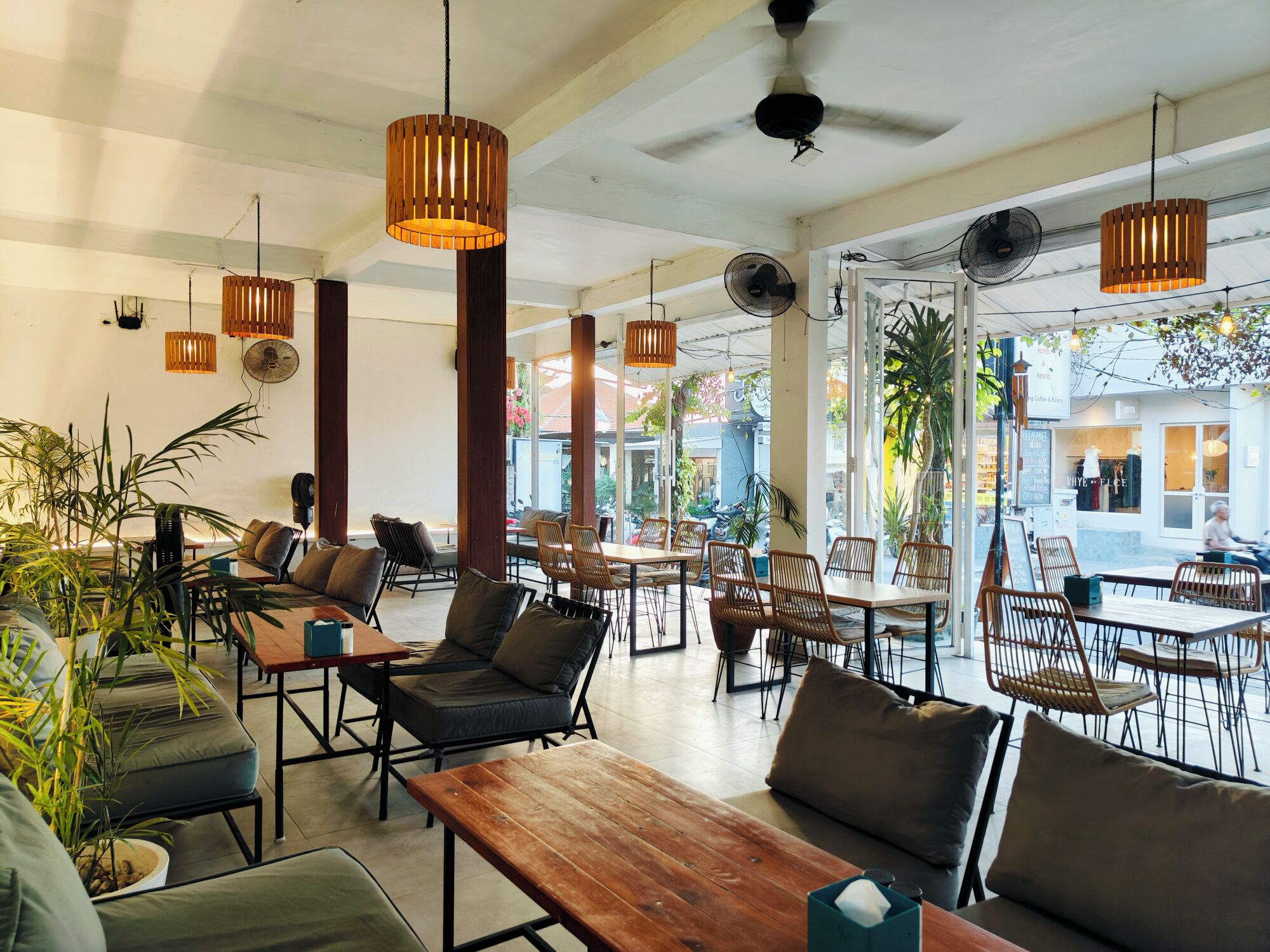 A spacious cafe with wooden tables and chairs, hanging pendant lights, and large windows offering views of the street.