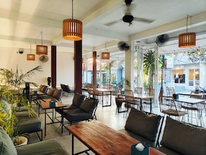 A spacious cafe with wooden tables and chairs, hanging pendant lights, and large windows offering views of the street. - Ashana Hotel Uluwatu (Pecatu)
