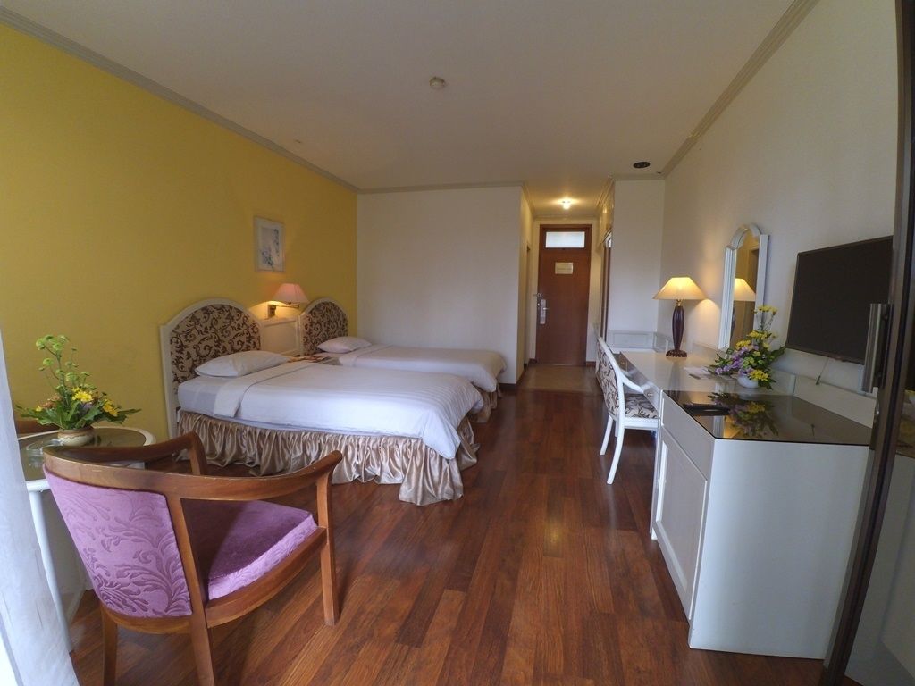 A hotel room with a wooden floor, a bed with a white bedspread, a desk with a computer, and a chair.