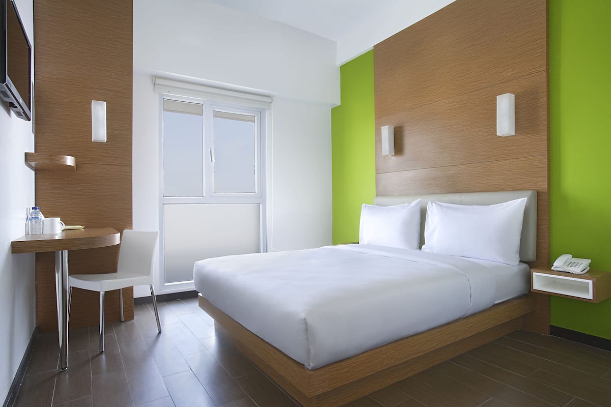 A modern hotel room with a large bed, a desk with a chair, a window, and a green accent wall.