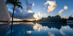 Ein klares Schwimmbecken spiegelt den Himmel und die umliegende Landschaft wider, darunter eine Palme und ferne Hügel.