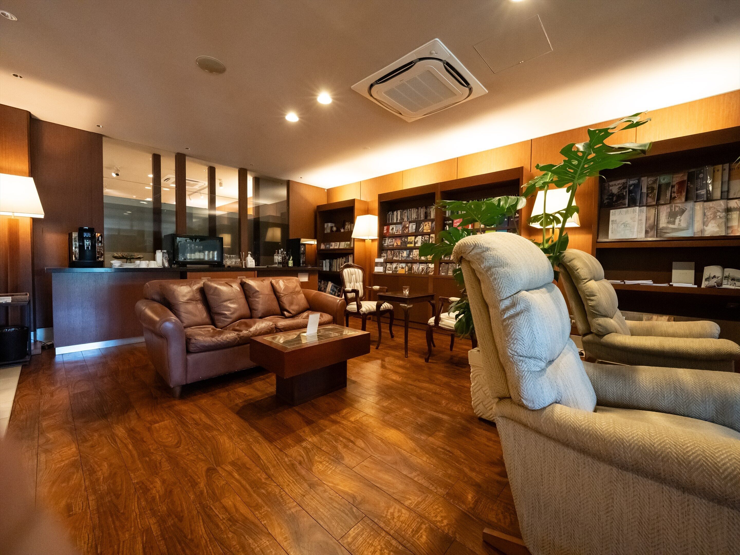 Una sala de estar moderna con un sofá de cuero, un sillón beige, una mesa de centro, una estantería y una planta grande.