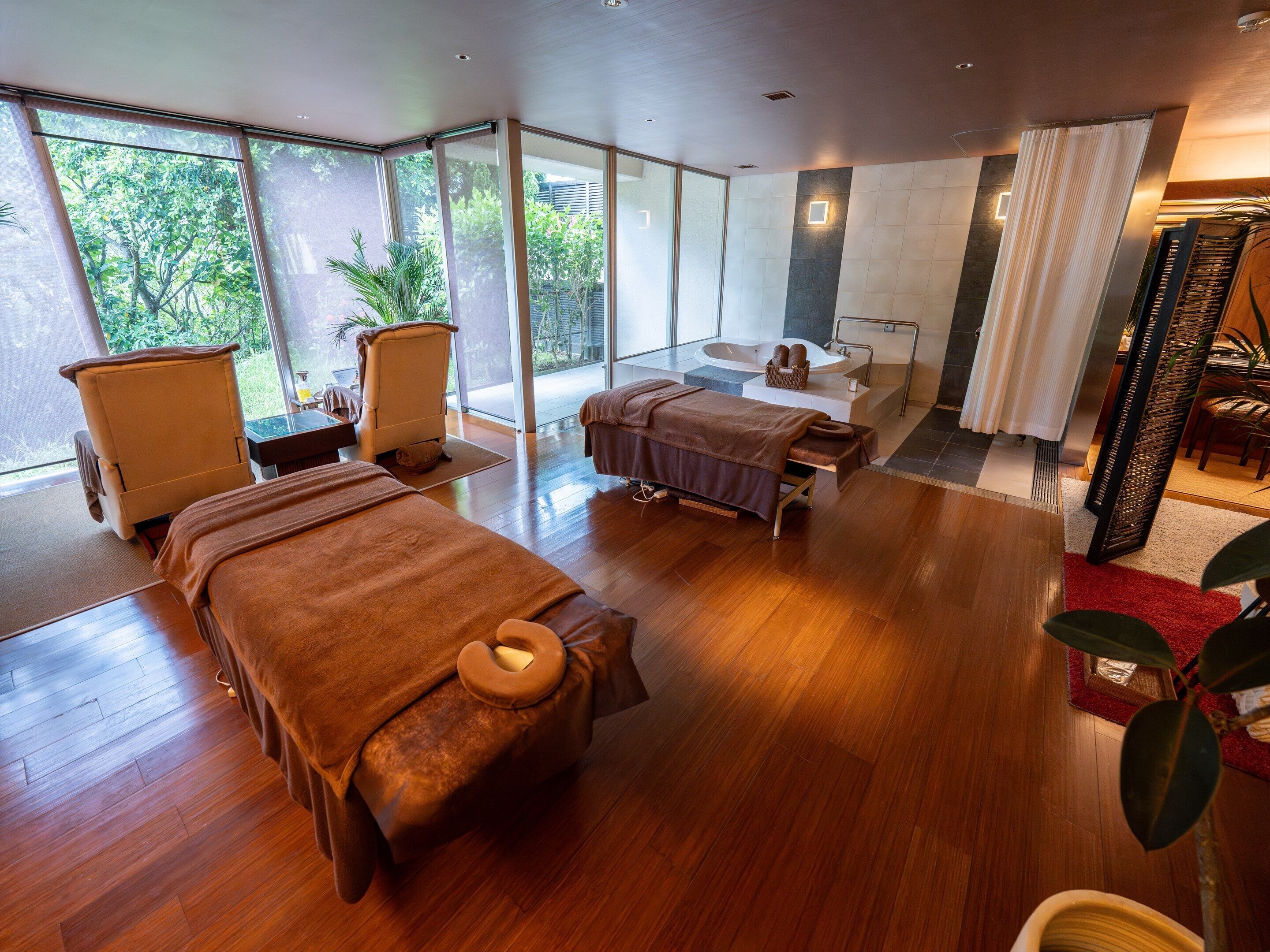 A modern spa room with two massage beds, a large window, and a bathtub.
