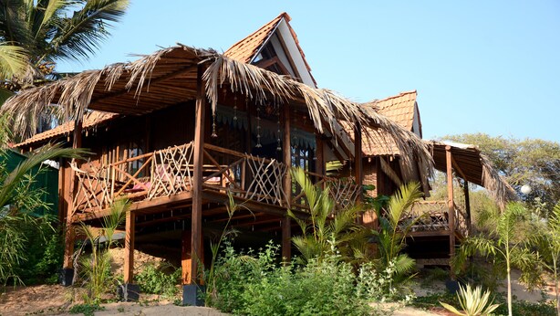 Una casa de madera con techo de paja, rodeada de follaje y palmeras.