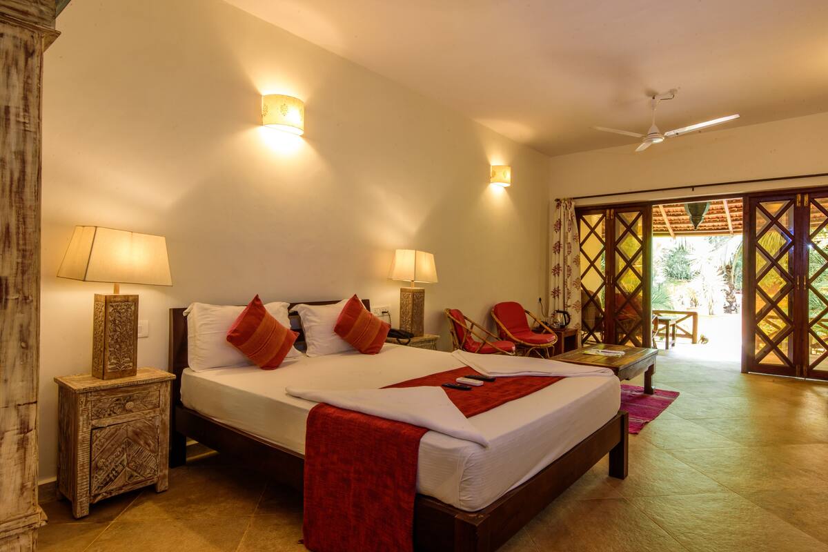 A hotel room with a large bed, two bedside lamps, a wooden nightstand, a red throw blanket, a wooden chair, and a view of the outdoors through a wooden door.