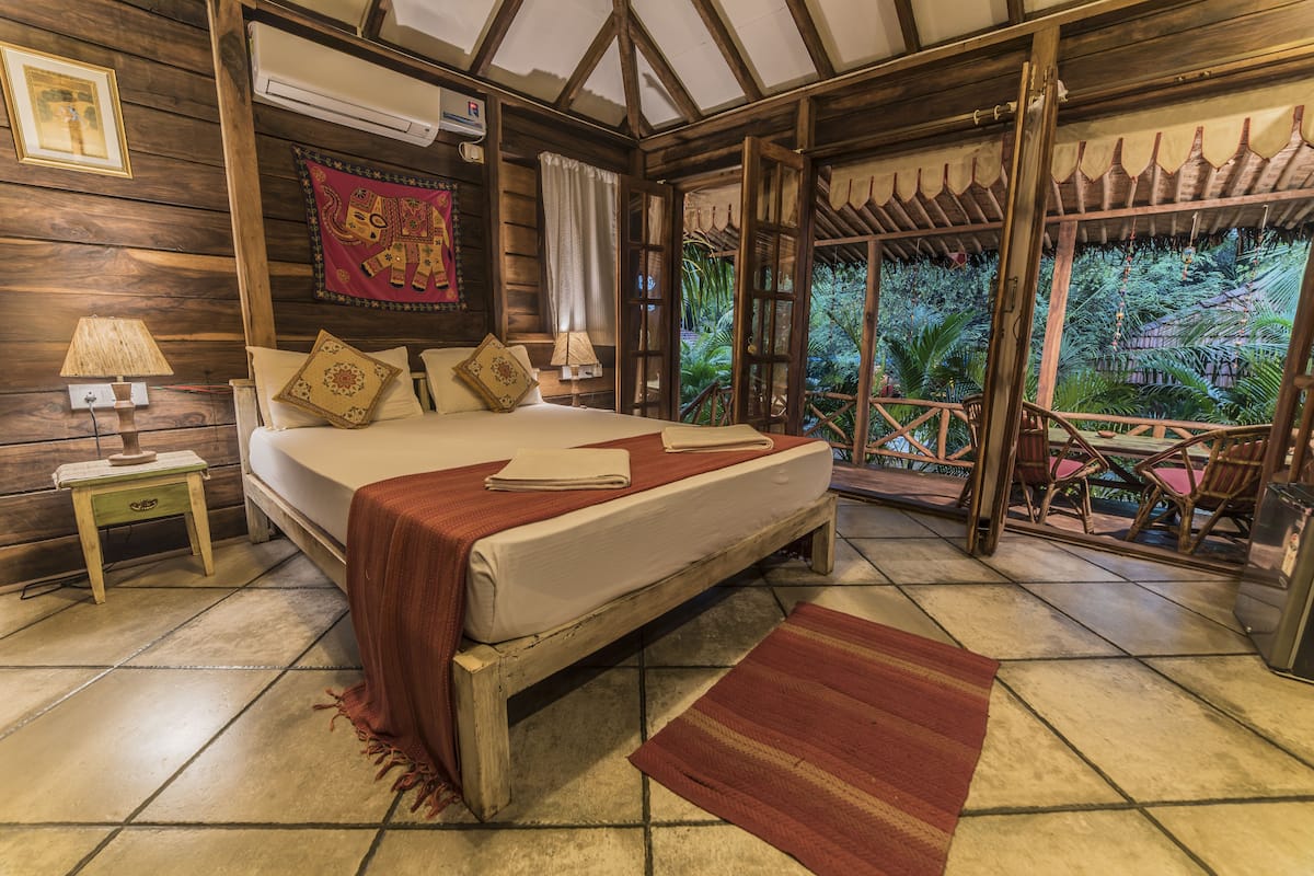 A wooden room with a bed, a nightstand, a window with curtains, and a view of greenery outside.