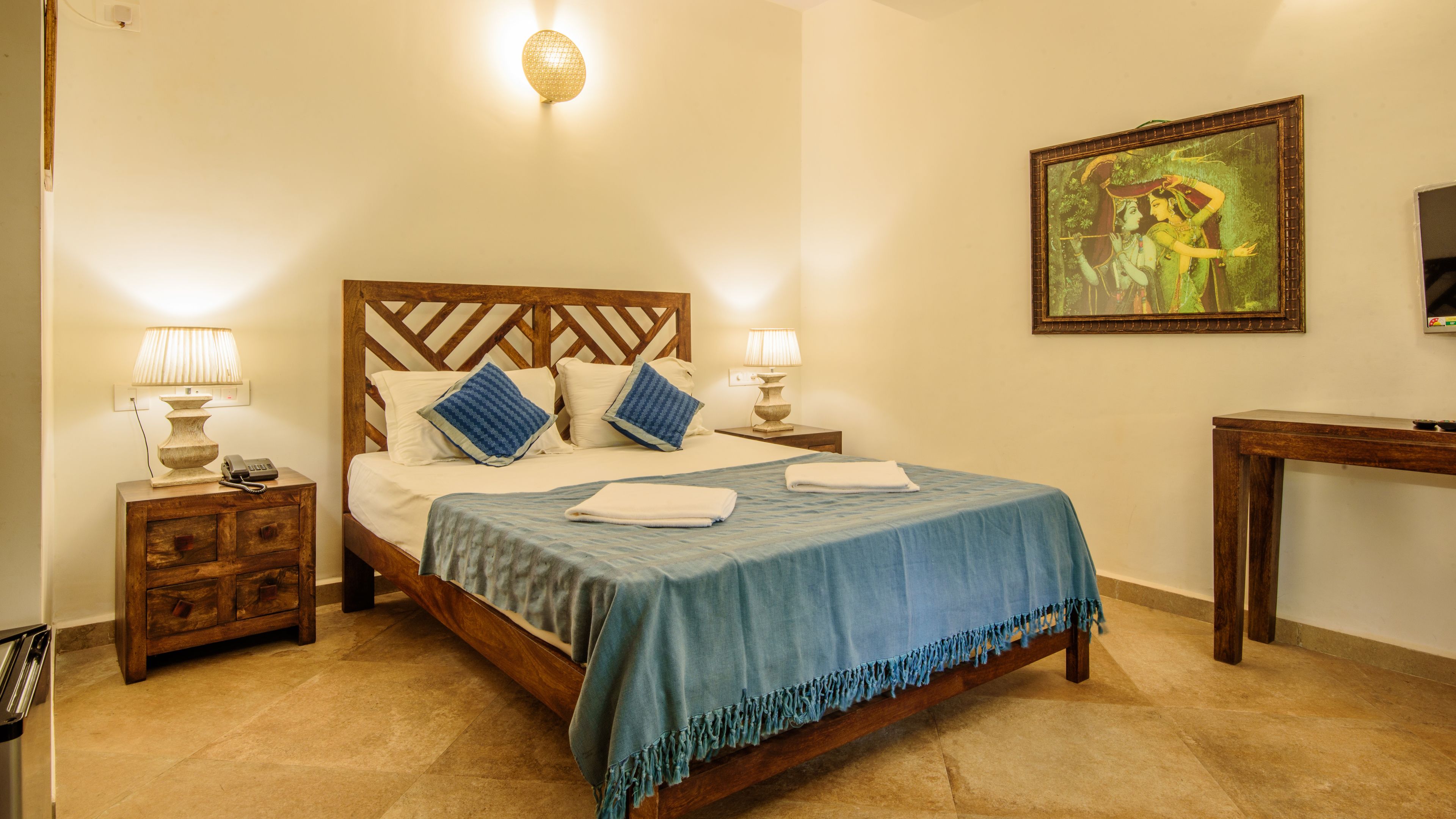A neatly arranged bedroom with a wooden bed, two bedside tables with lamps, a painting on the wall, and a wooden desk.