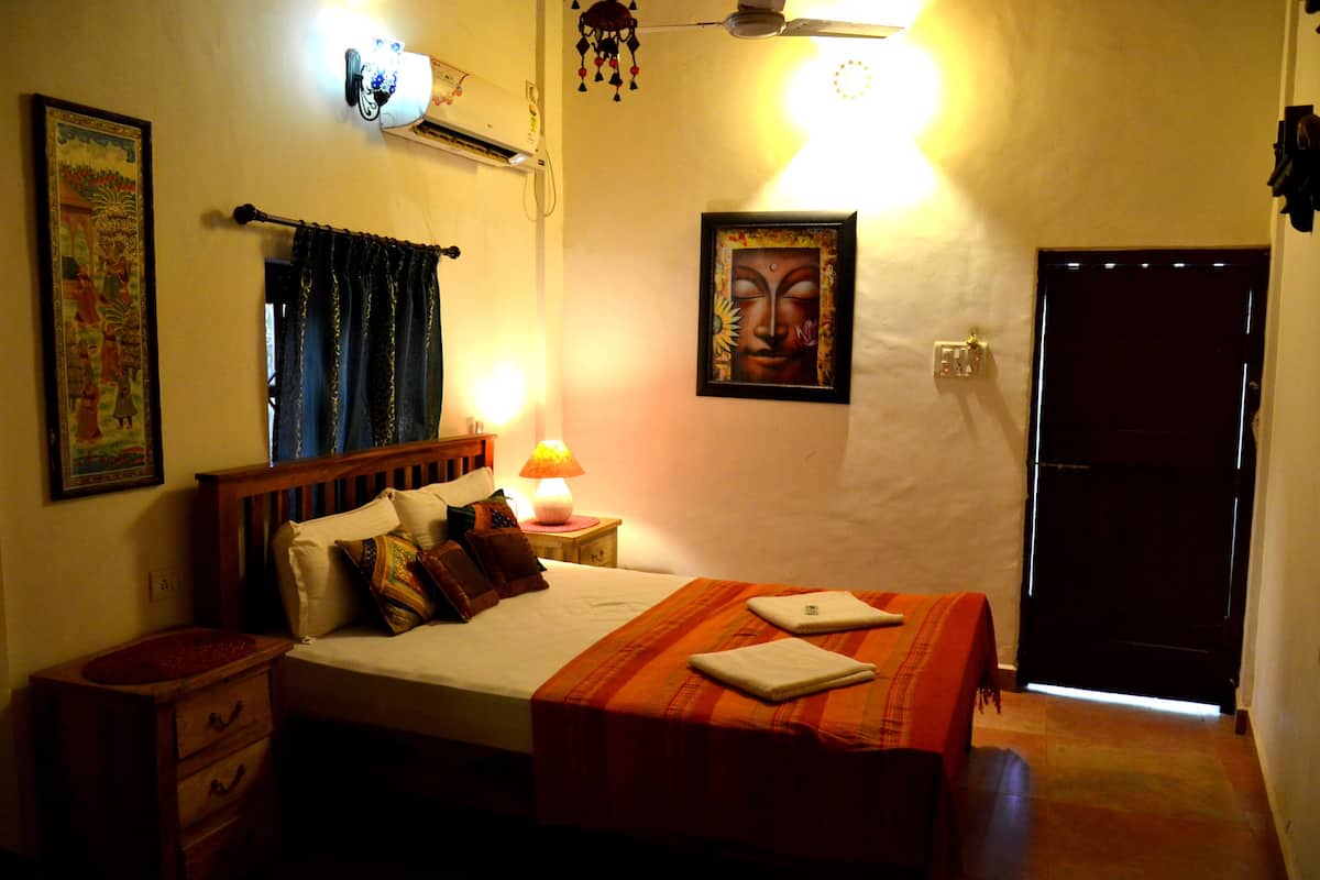 A hotel room with a bed, a nightstand, a wall-mounted air conditioner, a framed picture, and a door.