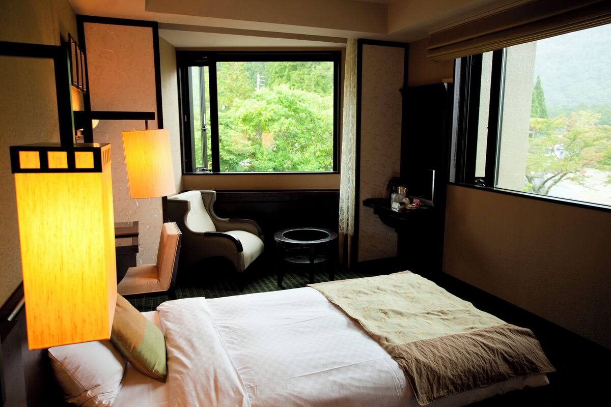 A hotel room with a bed, a desk, a chair, a lamp, a TV, and a window with a view of greenery.
