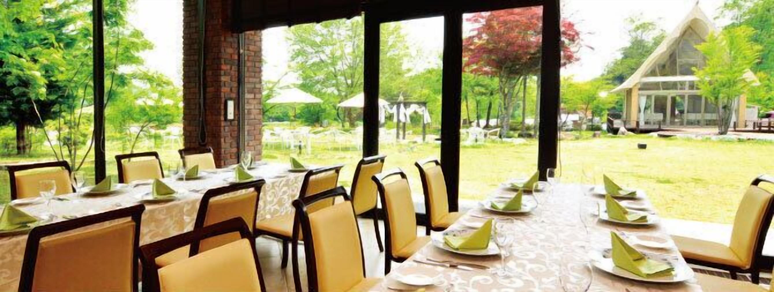 An outdoor dining area with tables set for a meal, surrounded by chairs, with a view of a building and trees in the background.
