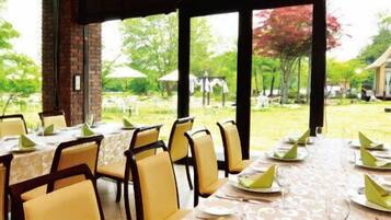An outdoor dining area with tables set for a meal, surrounded by chairs, with a view of a building and trees in the background.