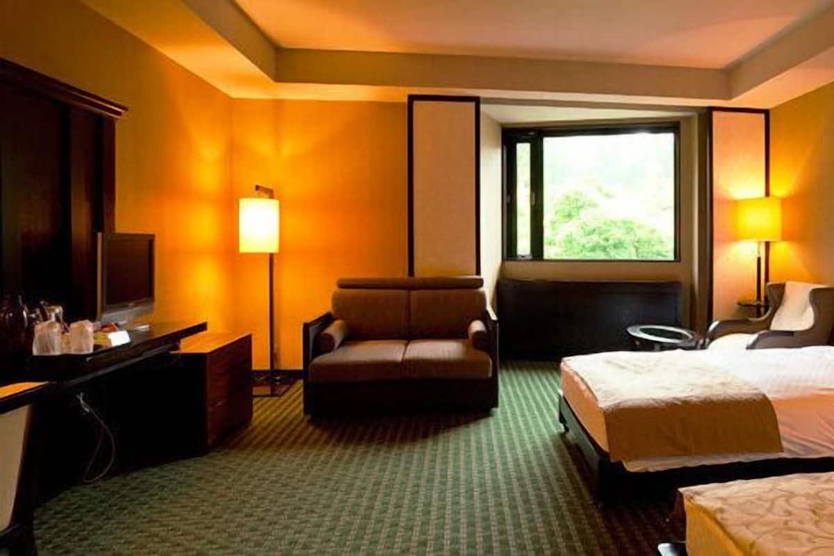 A hotel room with two beds, a desk with a chair, a sofa, and a window with a view of greenery.