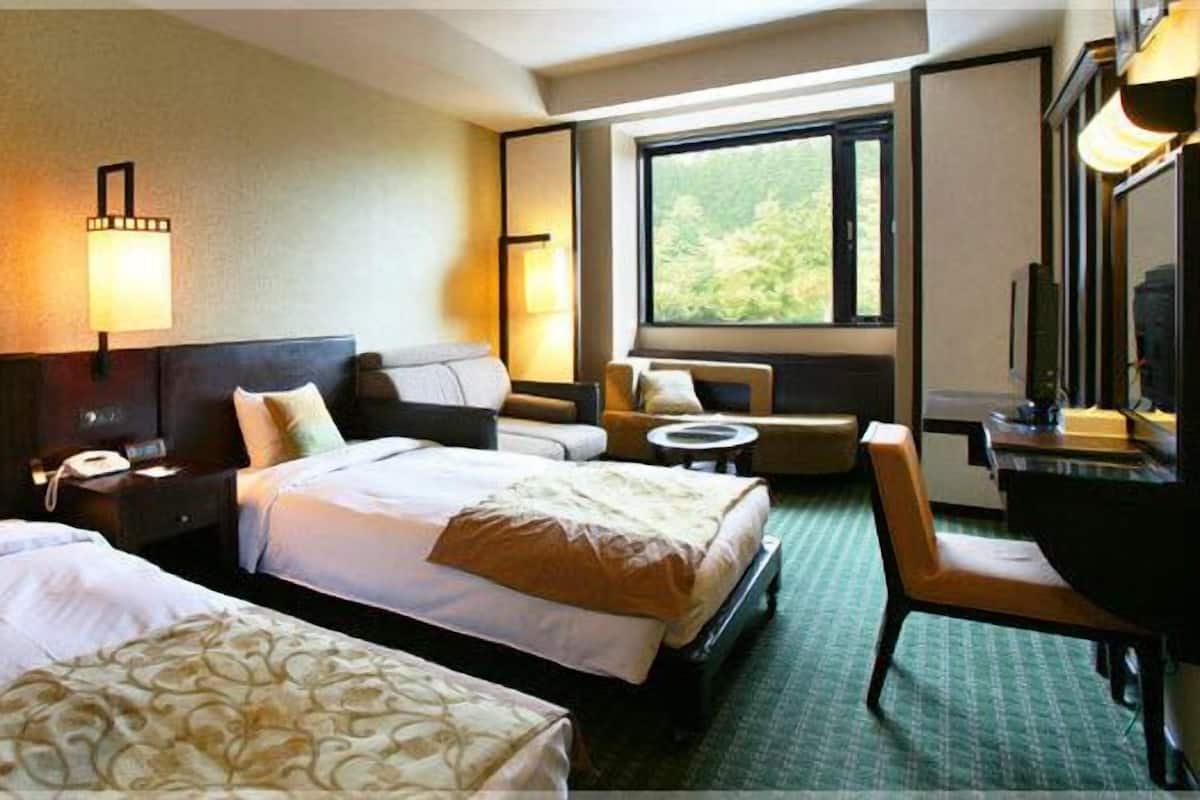 A hotel room with two beds, a desk, a chair, a small table, and a large window offering a view of greenery.