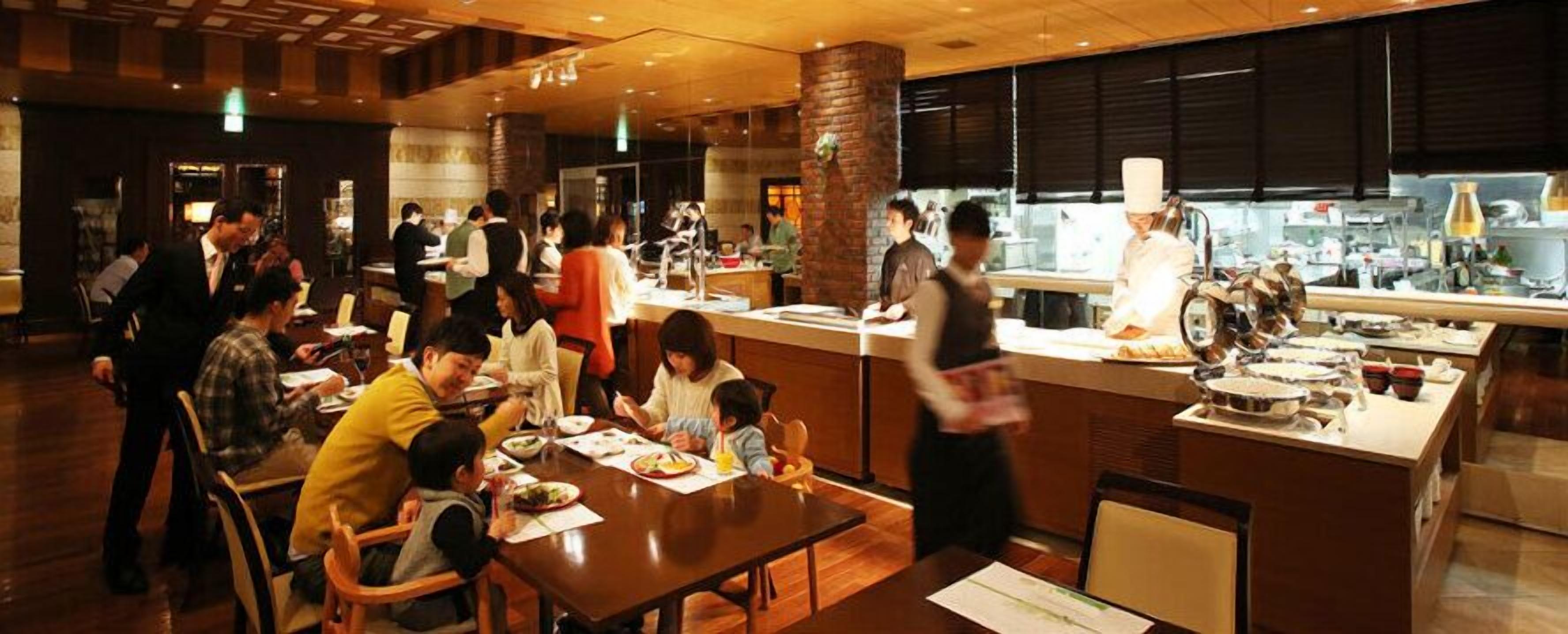 A restaurant with wooden tables and chairs, a kitchen area with chefs, and a counter for ordering.