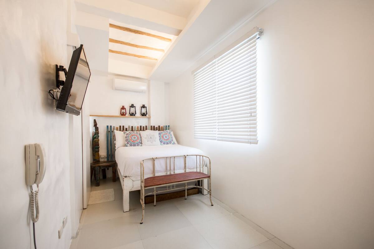 Une chambre à coucher comprenant un lit, un banc, un téléphone fixé au mur, une étagère avec des objets décoratifs et une fenêtre avec des stores.