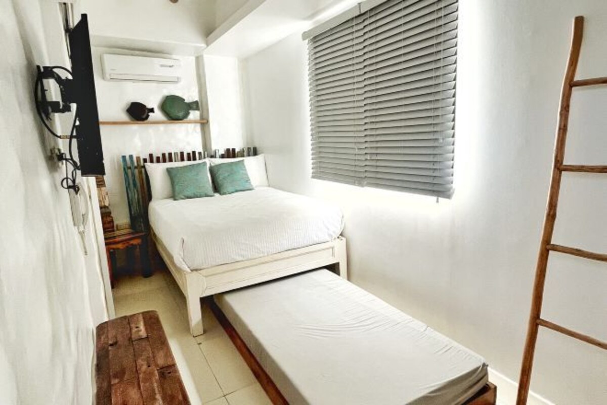 A small, compact bedroom with a single bed, a pull-out bench, a wooden ladder, and a window with blinds.