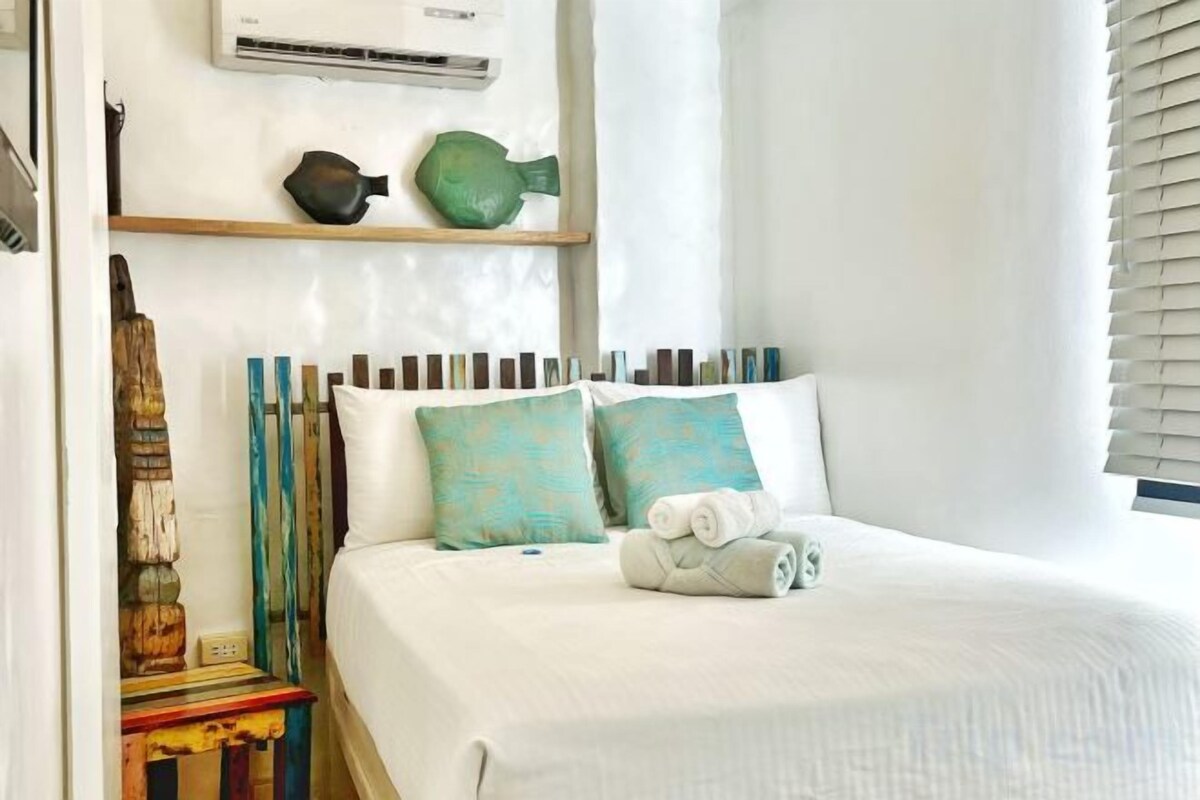 Een net opgemaakt bed met kussens en een opgerolde handdoek, een plank met decoratieve objecten en een wandairconditioner.