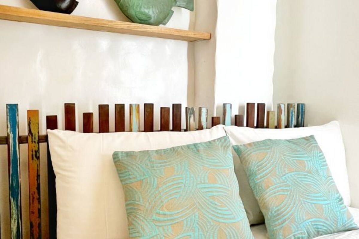 Een bed met een wit hoofdeinde en twee gestreepte kussens, een houten plank erboven, en een rij boeken aan de muur.