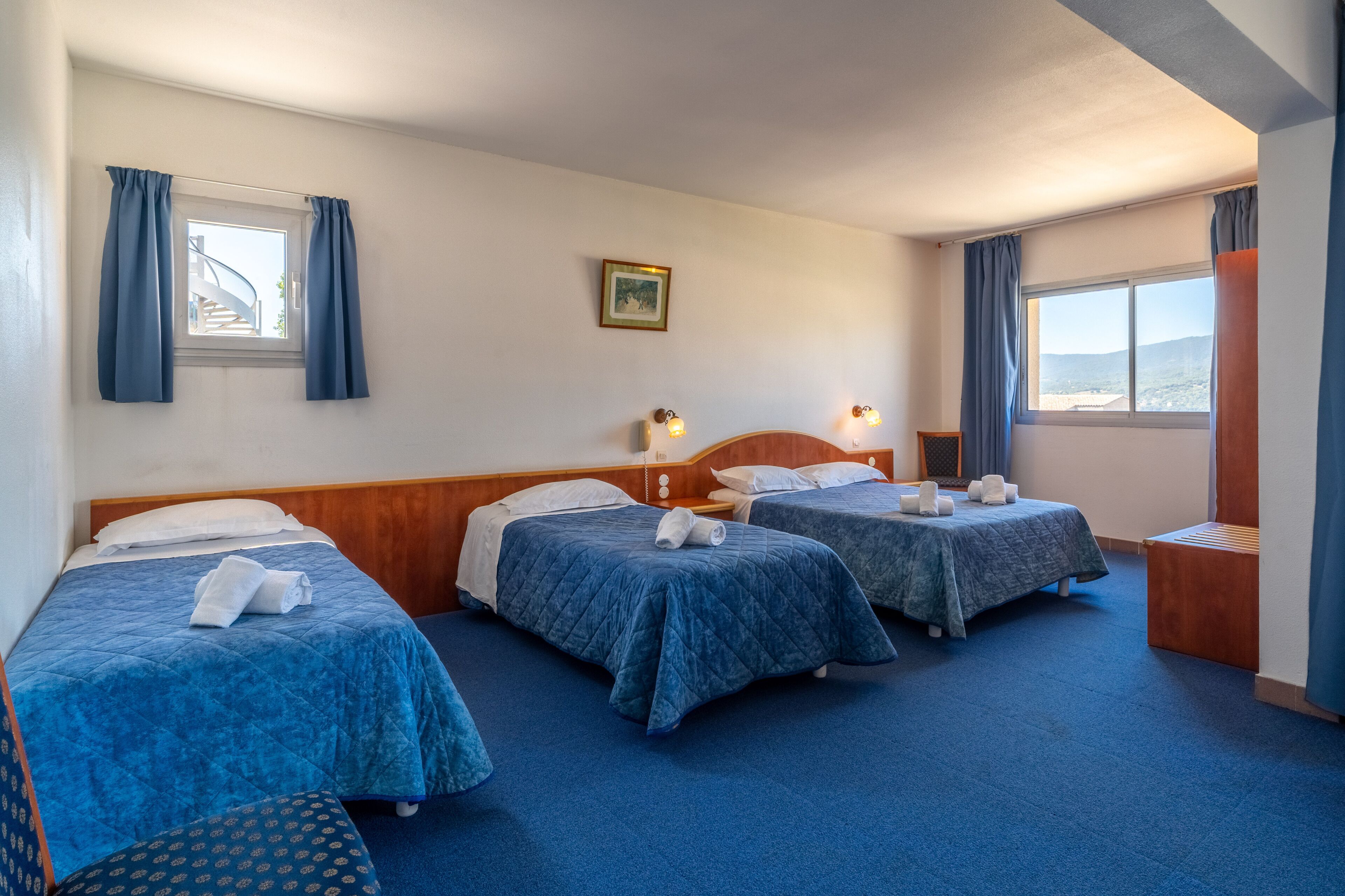 Una habitación de hotel con tres camas, cabecera de madera, una mesita y una ventana con cortinas.