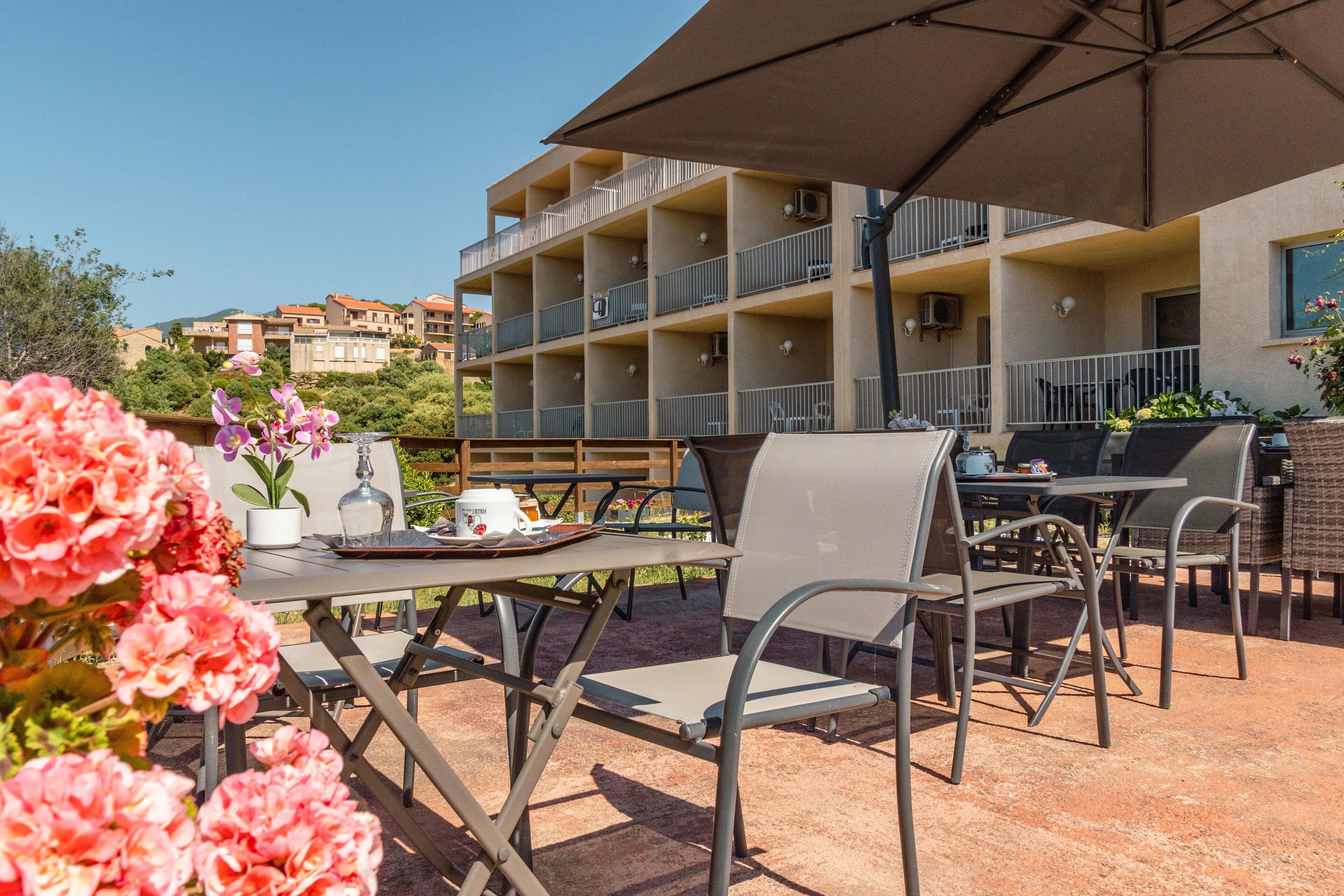 An outdoor patio with a table and chairs, a large umbrella, and a view of a multi-story building with balconies.