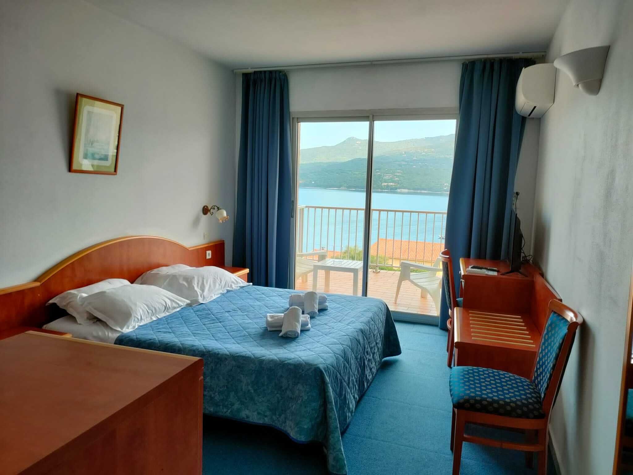 A hotel room with a bed, a desk, a chair, and a balcony with a view of the sea.