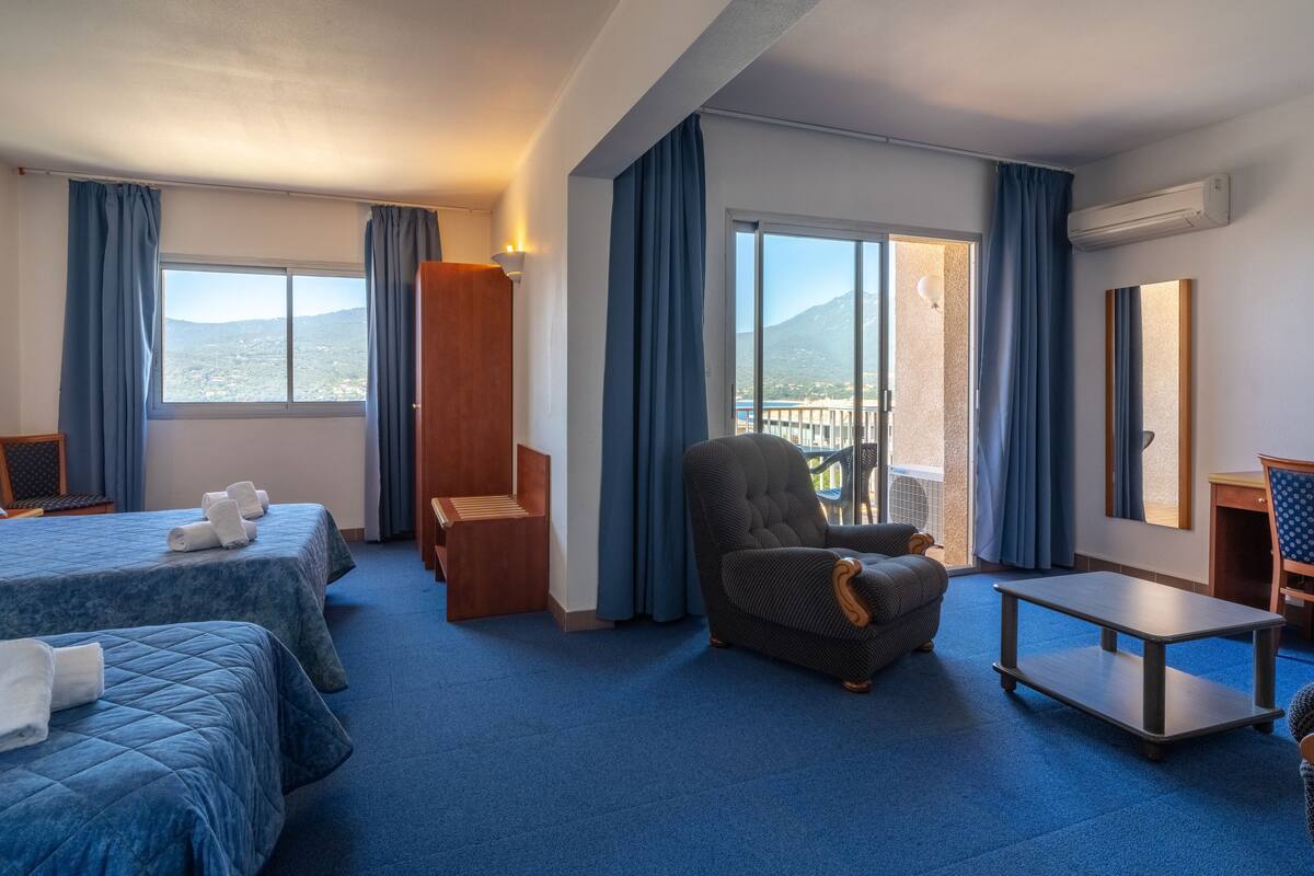 Une chambre d’hôtel avec deux lits, une chaise, une table et un balcon offrant une vue sur les montagnes.