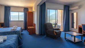A hotel room with two beds, a chair, a table, and a balcony with a view of the mountains.