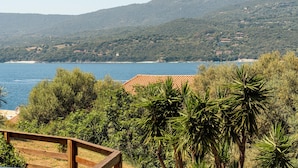 A coastal landscape with a wooden deck, dense greenery, and a view of the sea and hills.