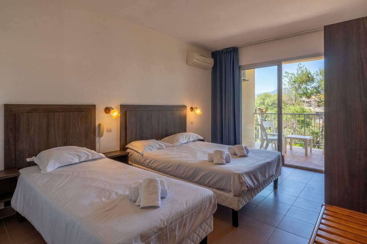 Une chambre d’hôtel avec deux lits, une tête de lit en bois, un téléphone sur la table de chevet et un balcon avec une table et des chaises.