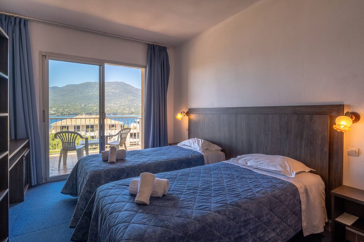 Une chambre d’hôtel avec deux lits, un balcon offrant une vue sur la mer et les montagnes, et une tête de lit en bois.