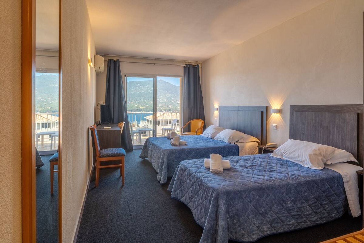 Une chambre d’hôtel avec deux lits, un bureau, une chaise et un balcon offrant une vue sur les montagnes.