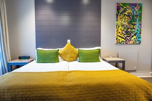 A hotel room with a bed, green and yellow pillows, a nightstand, a painting, and a radiator. - Beachwalk Bed and Breakfast (Gqeberha (Port Elizabeth))