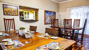 Un coin repas avec une table en bois dressée pour un repas, entourée de chaises, des œuvres d’art ornant les murs et une fenêtre agrémentée de rideaux.