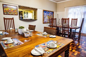 Un comedor con una mesa de madera preparada para una comida, rodeada de sillas, con obras de arte en las paredes y una ventana con cortinas.