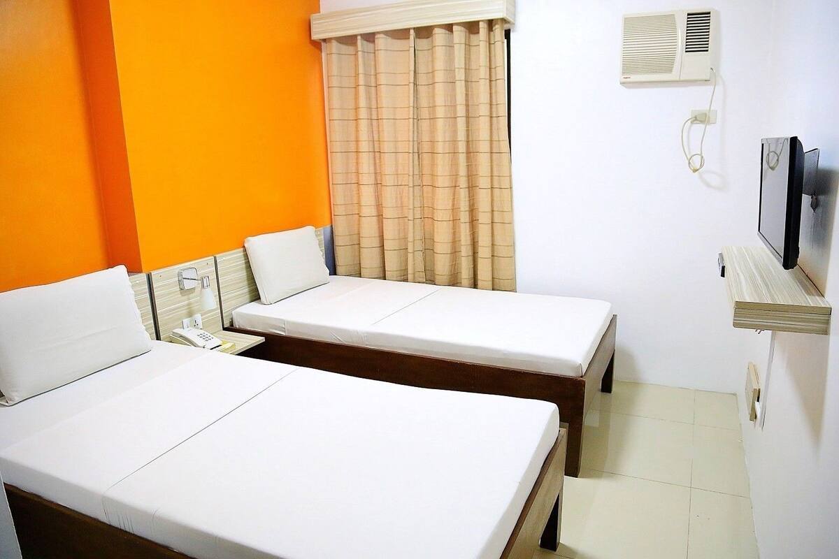 Quarto de hotel com duas camas de solteiro, uma mesa de cabeceira com telefone, televisão na parede e uma janela com cortinas.