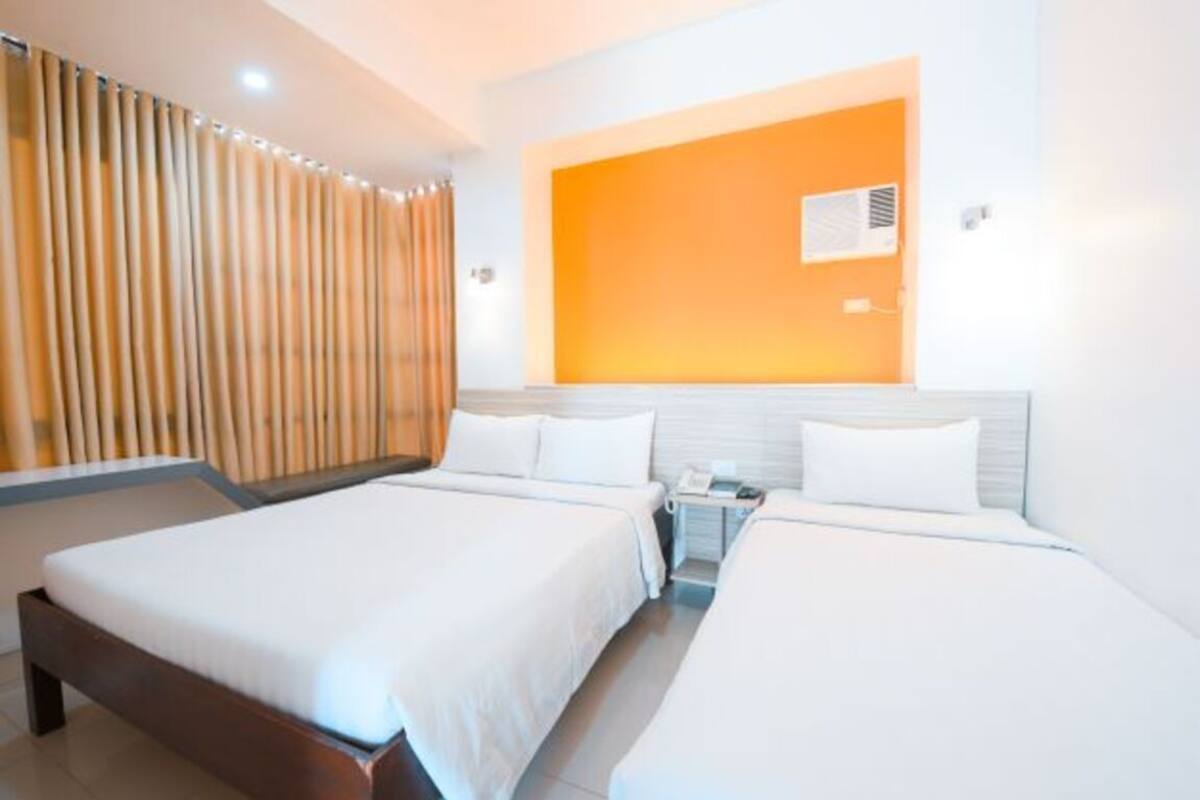 Quarto de hotel com duas camas, cortina de madeira, parede de destaque laranja e uma pequena mesa de cabeceira com telefone.