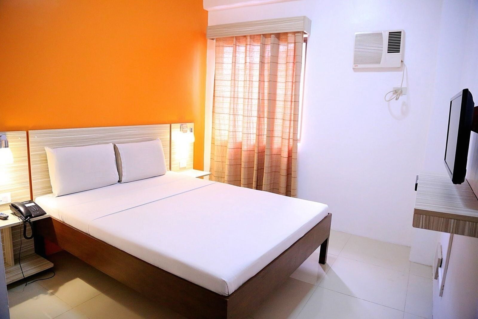 A hotel room with a bed, bedside lamps, a telephone, an air conditioning unit, and a television.