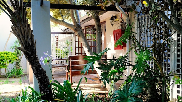 Un jardín con un banco de madera, un pilar blanco y una ventana con contraventanas rojas.