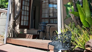 Un espacio al aire libre techado con mesa y sillas, rodeado de plantas tropicales y un edificio con amplias ventanas.