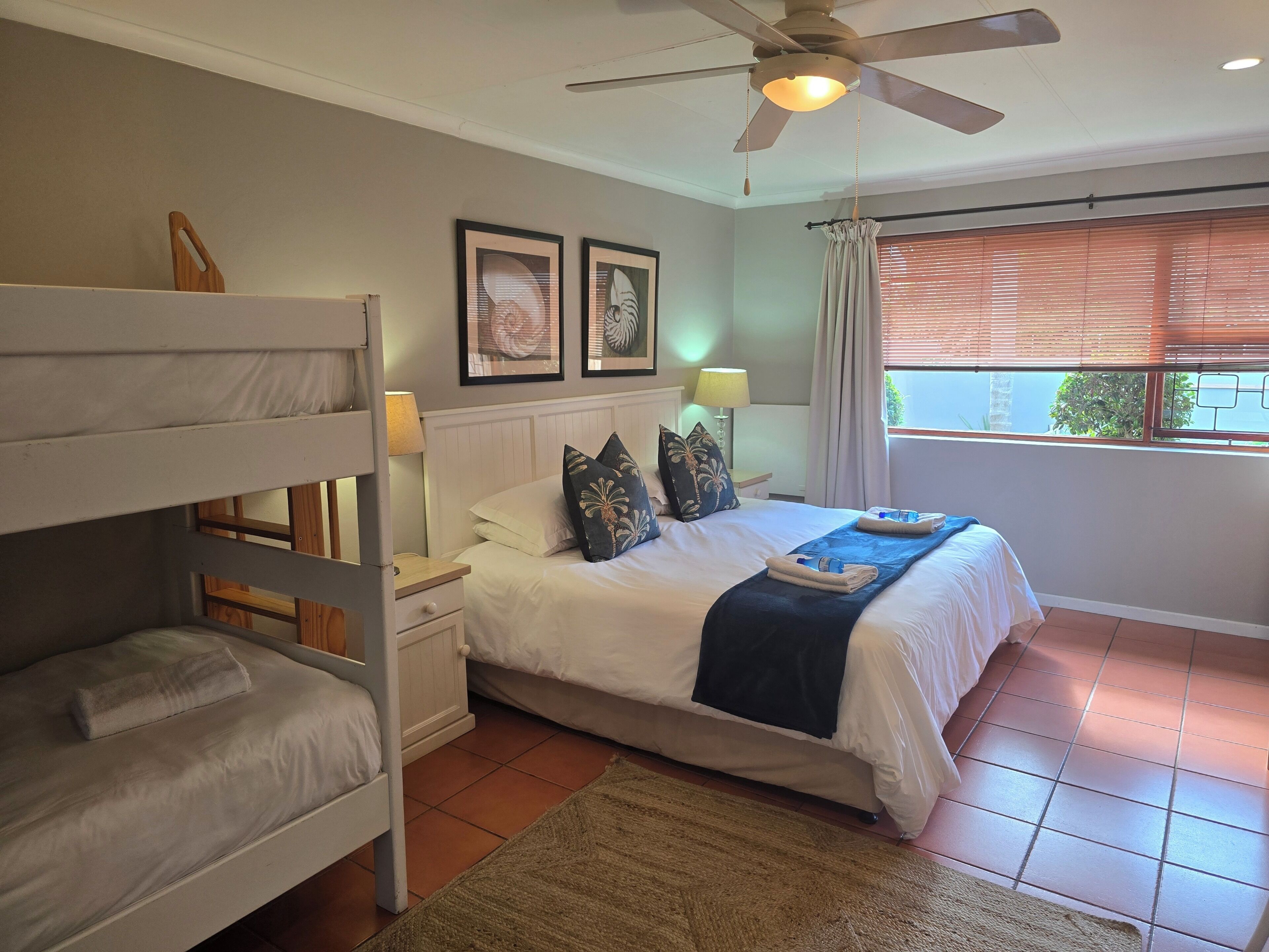 A bedroom with bunk beds, a bed with pillows, a bedside table with a lamp, and a window with blinds.