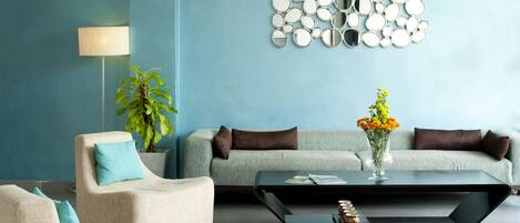 A modern living room with a blue wall, a round mirror wall art piece, a floor lamp, a potted plant, a sofa, two armchairs, and a coffee table with a vase of flowers.