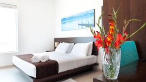 A neatly made bed with white and brown bedding, a vase with red flowers, and a view of a dock and mountains through a window. - The Beach Apartments (Negombo)