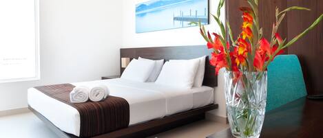 Una cama bien tendida con ropa de cama blanca y marrón, un jarrón con flores rojas, y una vista de un muelle y montañas a través de una ventana.