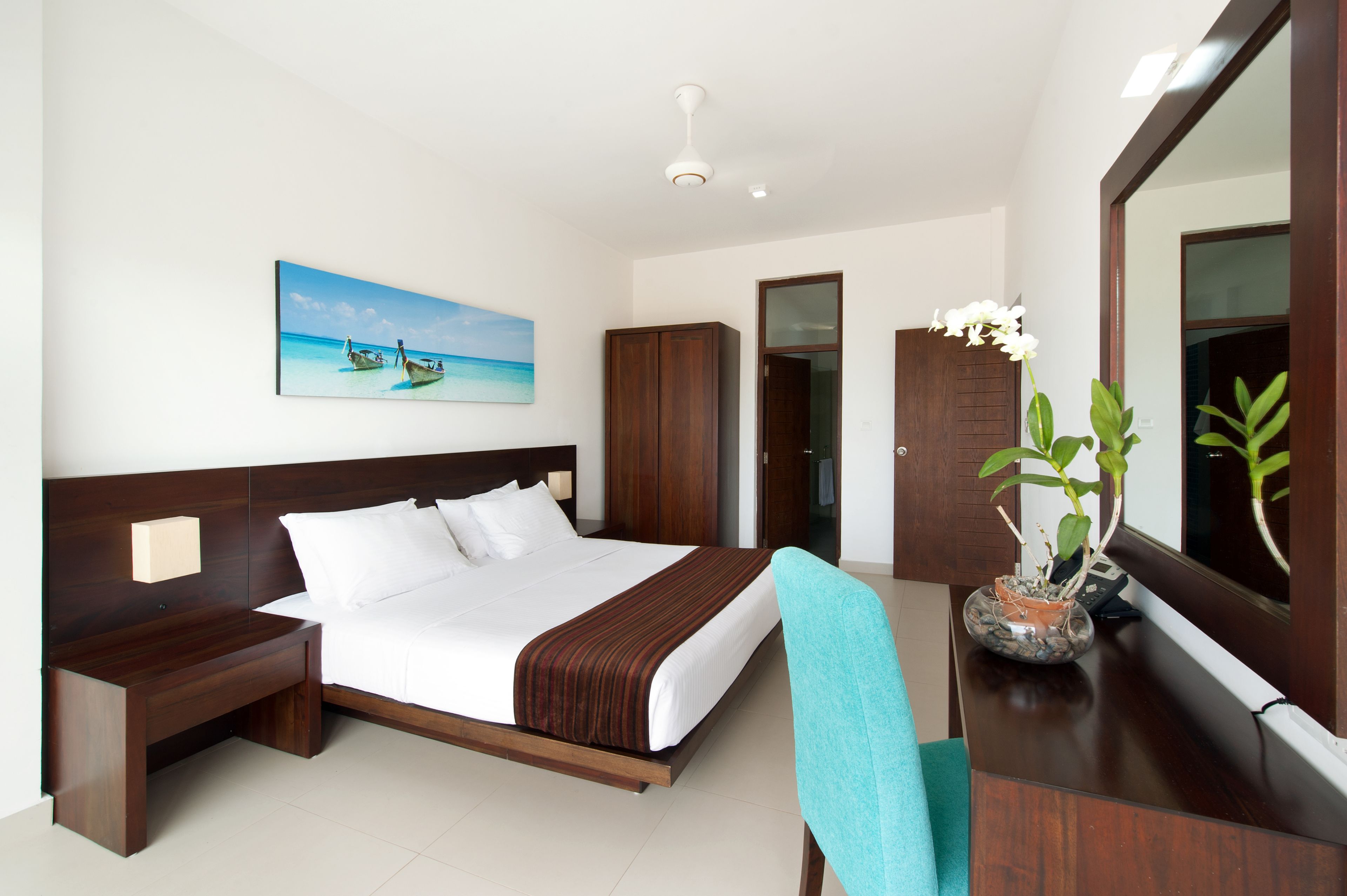 A hotel room with a large bed, wooden headboard, a nightstand, a desk with a chair, a potted plant, and a painting of a beach scene.