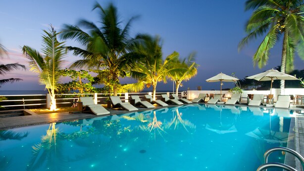 A poolside area with lounge chairs and umbrellas, palm trees, and a view of the ocean. - The Beach Apartments (Negombo)