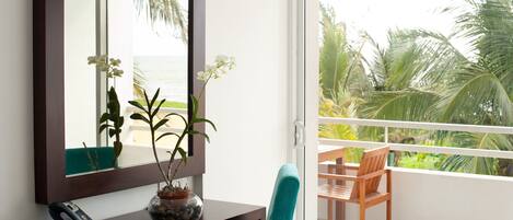 A room with a large mirror, a wooden desk with a vase of flowers, a telephone, and a view of palm trees through a balcony door.