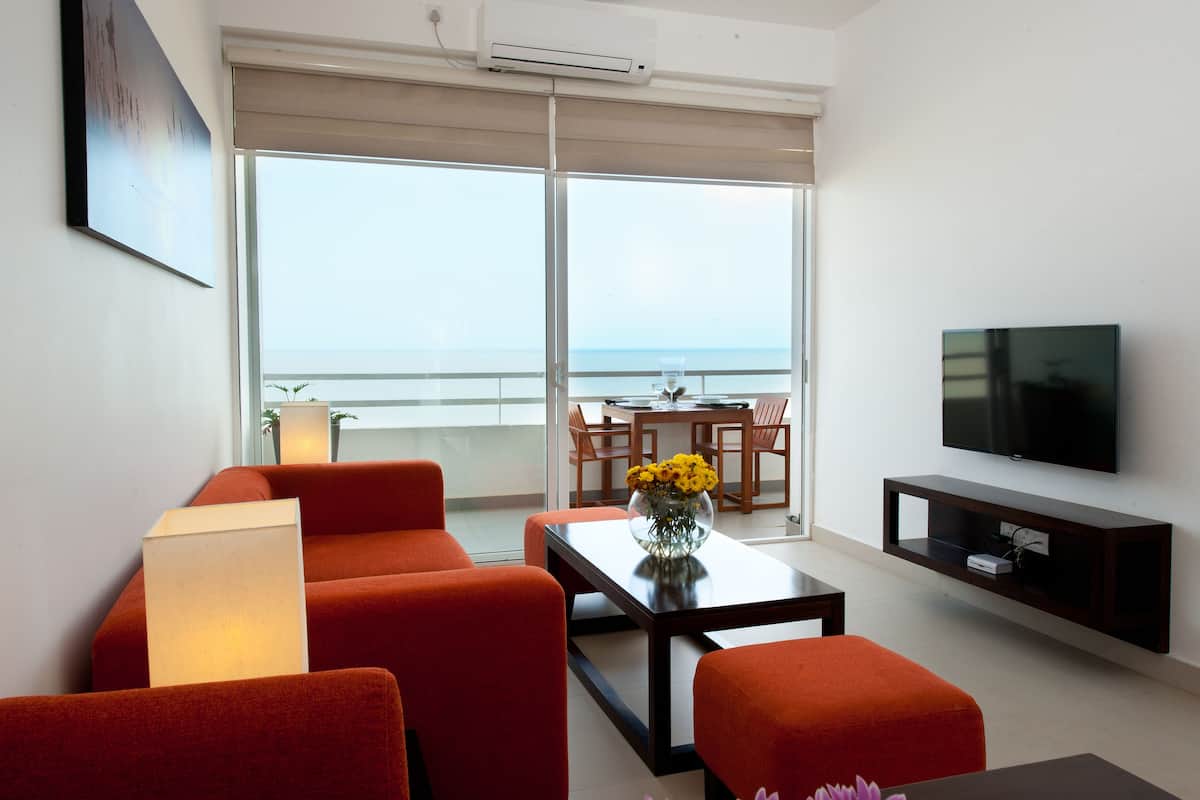 A modern living room with a sofa, a coffee table with a vase of flowers, a flat-screen TV, and a balcony with a view of the sea.