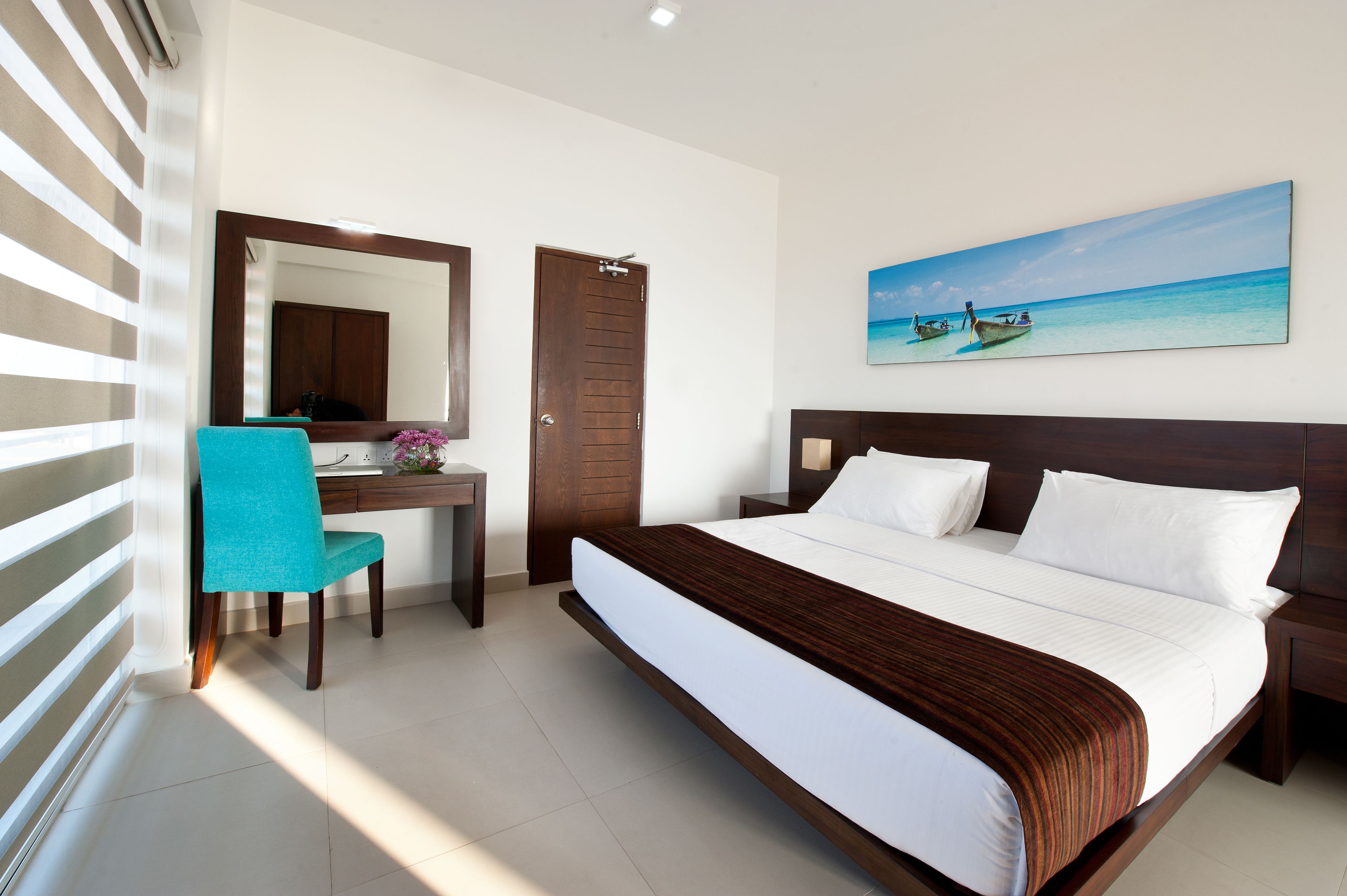 A hotel room with a large bed, a desk with a chair, a mirror, and a painting of a beach scene.